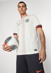 Maglia da football Nike bianca con logo circolare, accenti a strisce verdi e rosse sulle maniche e linee verticali rosse e verdi. Palla a motivi bianchi e neri.