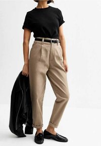 T-shirt en coton noir, pantalon beige taille haute, ceinture noire, mocassins noirs. Veste légère drapée sur le bras gauche. Fond neutre.