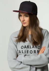 Sort kasket med flad skygge, med en farverig ternet inderside, båret med en grå sweatshirt, der viser "OAKLAND CALIFORNIA" med fede bogstaver.