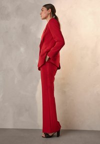 Rød skræddersyet blazer og matchende bukser lavet af glat stof. Blazeren har rene linjer, og bukserne har side-striber, parret med sorte hæle.