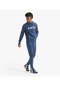 Felpa blu navy con logo bianco, cordino e tasca a marsupio, abbinata a pantaloni della tuta coordinati e scarpe da ginnastica multicolori.