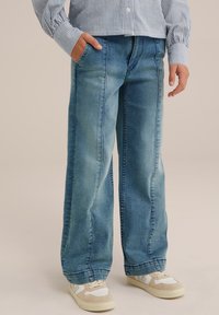 Široke modre kavbojke z zbledelim videzom, dvema stranskima žepoma in ravno silhueto so kombinirane z bež supergami.