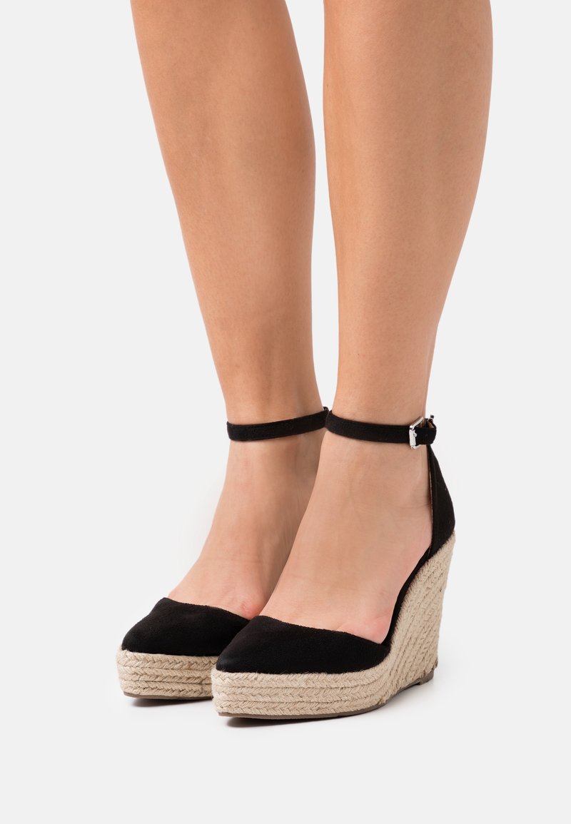 Chaussures compensées noires à bout pointu et bride à la cheville. Confectionnées en tissu doux avec une plateforme en jute tissé. Agrémentées d'une boucle argentée.