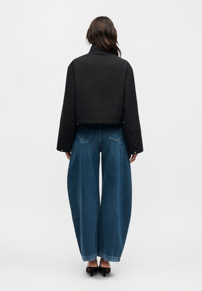 Γυναίκα με πλάτη, φοράει μαύρο κοντό μπουφάν, μπλε τζιν παντελόνι με φαρδιά ποδόγυρα και μαύρα παπούτσια με τακούνι σε απλό λευκό φόντο.