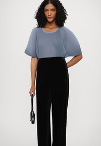 Haut plissé bleu clair avec des manches courtes et larges, associé à un pantalon fluide noir. La mannequin tient un petit sac à main noir avec une bandoulière.