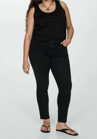 Femme portant un haut noir sans manches, un jean skinny noir, un collier en forme d'étoile de mer et des tongs noires, debout avec une main dans la poche.