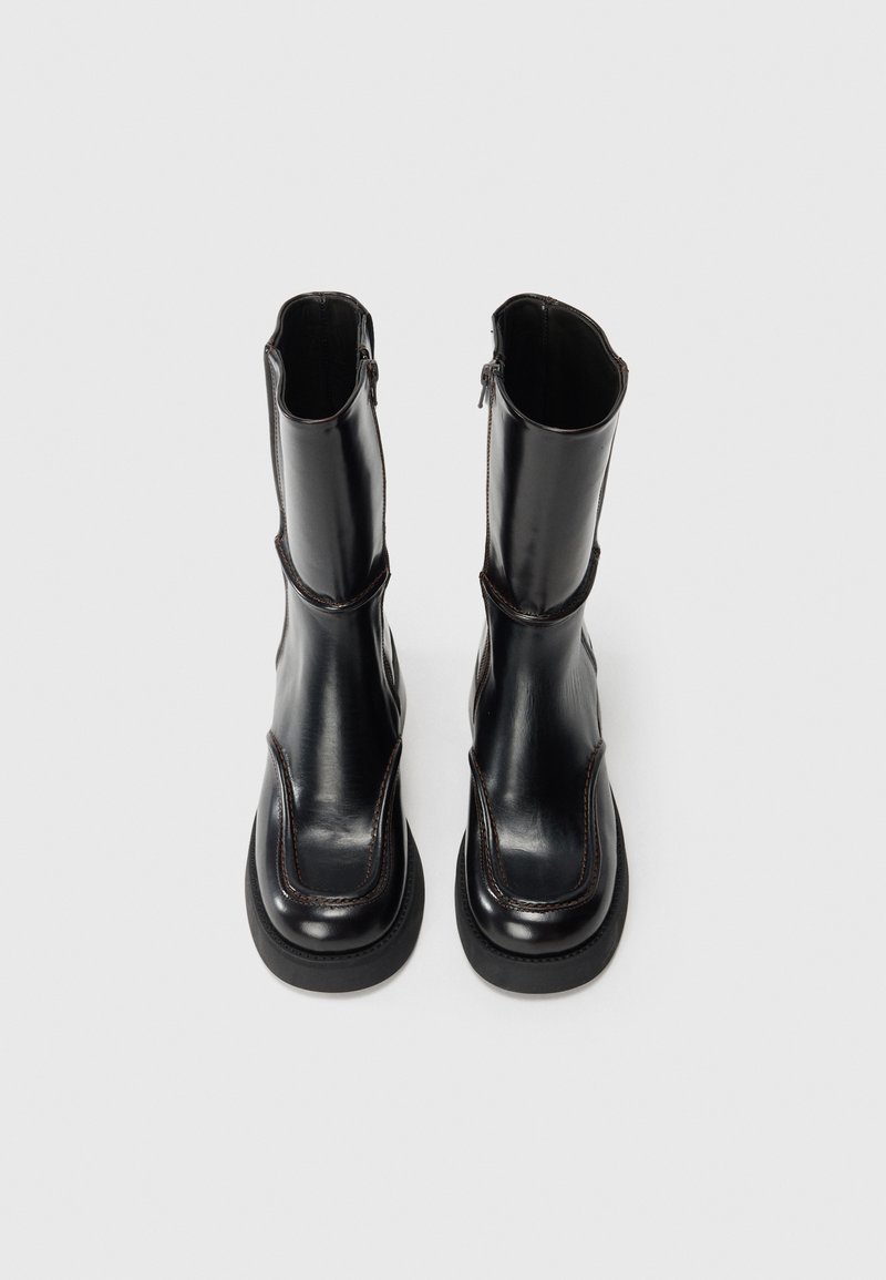 Une paire de bottes en cuir noir mi-mollet avec des bouts carrés, des semelles épaisses et des fermetures éclair sur les côtés, vues de dessus sur un fond blanc uni.