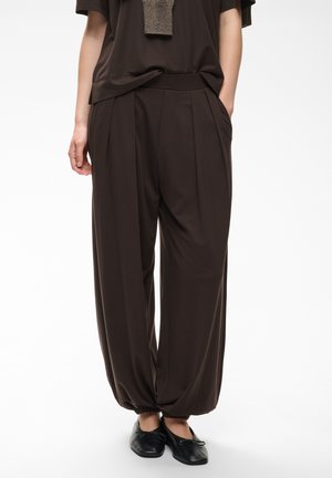 Persona con pantalones marrones plisados y sueltos con puños elásticos combinados con bailarinas negras, de pie con las manos en los bolsillos.