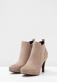 Un par de botas de ante beige hasta el tobillo con cremalleras laterales y tacones altos sobre una superficie y fondo blancos.
