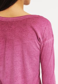 Pitkähihaisen yläosa liukuvärissä marjaisaa sävyä, pyöreällä pääntiellä ja pehmeällä, teksturoidulla kankaalla, jossa on hienovarainen kiilto.