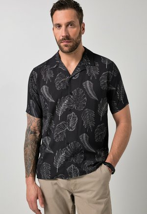 Mann mit Bart, der ein schwarzes kurzärmeliges Hemd mit grauen Blattmustern und beige Hose trägt, steht mit einer Hand in der Tasche.