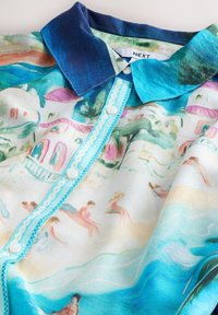 Chemise avec col bleu et patte de boutons, présentant une scène de plage style aquarelle avec des personnes, des parasols et des vagues de l’océan en couleurs pastel.