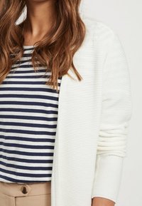 Vit ribbad cardigan över en marinblå och vit randig skjorta, med en avslappnad passform och mjuk textur. Bruna byxor med synlig knappdekoration framtill.