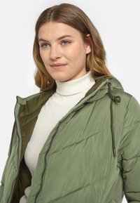 Gelegenheitsgrüne gefütterte Jacke mit Kapuze, ausgestattet mit einem Reißverschluss und einem schrägen gesteppten Muster. Getragen über einem weißen gerippten Rollkragen.