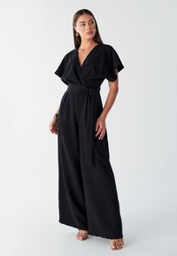 Črn ovit kombinezon s kratkimi rokavi z rahlim valom in z zavezovanjem v pasu. Ima globok V-izrez in široke hlačne nogavice. Gladek tekstura.