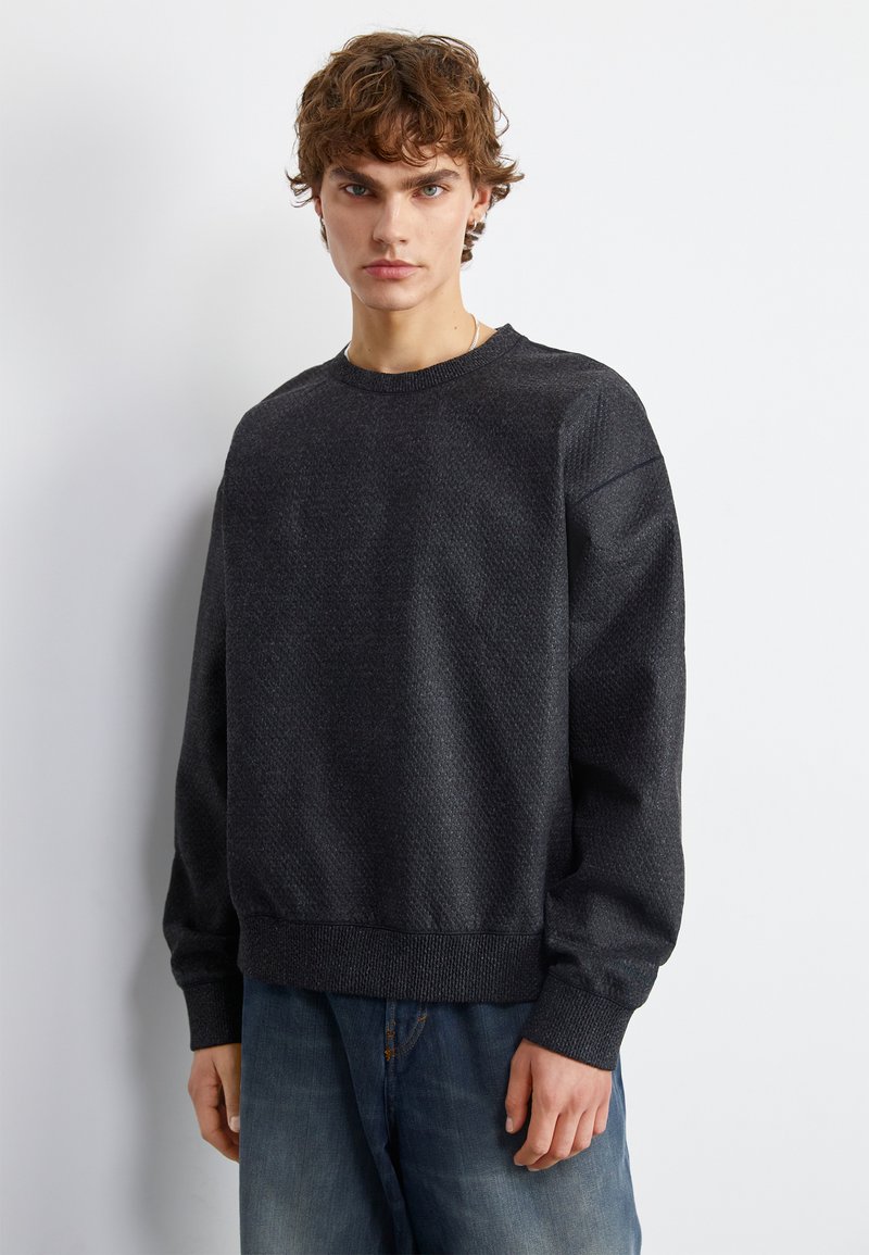 Ciemnoszary, teksturowany sweatshirt z okrągłym dekoltem i prążkowanymi mankietami, w połączeniu z jasnoniebieskimi, luźnymi dżinsami, na jednolitym białym tle.