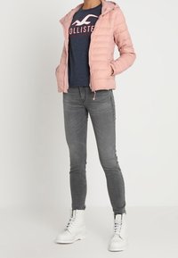 Jasnoróżowa pikowana kurtka z kapturem, zapinana na zamek, z przeszywanym wzorem. Połączona z granatowym T-shirtem i szarymi dżinsami, noszona z białymi butami.