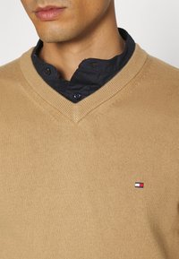 Beżowy sweter z dekoltem w serek wykonany z dzianiny. Posiada małą naszywkę z logo w kolorach czerwonym, białym i niebieskim po lewej stronie. Nałożony na czarną koszulę z kołnierzykiem.