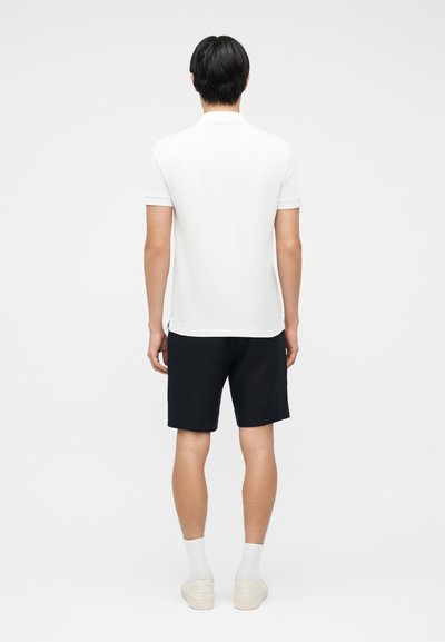 Άνδρας που φοράει λευκό πόλο με κοντά μανίκια, μαύρο σορτς, λευκές κάλτσες και λευκά αθλητικά παπούτσια, όρθιος με την πλάτη προς τα εμπρός σε απλό λευκό φόντο.