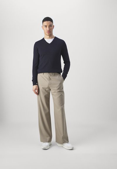 Pull en V bleu marine, pantalon beige à jambes larges et baskets blanches. Tissu lisse, coupe décontractée et détails de couture visibles.