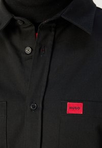 Chemise noire à boutons avec col, boutons visibles, poche poitrine avec un patch rouge portant la mention « HUGO », portée sur un col roulé noir.