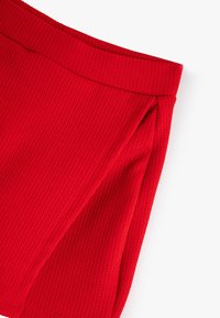 Close-up van een rode, gestructureerde stof rokhoek met een gevouwen detail en tailleband op een witte achtergrond.