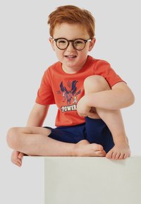 Rotes Baumwoll-T-Shirt mit einem Grafikdruck, kombiniert mit marineblauen Shorts. Kind mit Brille sitzt barfuß auf einer weißen Plattform und lächelt.