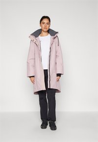 Parka impermeabile rosa con cappuccio, dettagli neri e chiusura con zip. Indossata sopra una camicia bianca, abbinata a pantaloni neri e stivali.