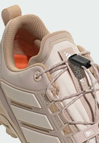 Jasno beżowe buty sportowe z siateczkową cholewką, plastikowymi akcentami, teksturowanymi sznurówkami i wzmocnionym językiem pięty. Jasnopomarańczowe wykończenie wnętrza.