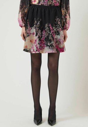 Kratka črna cvetlična krilo z rožami v roza, bež in vijolični barvi, nošeno s prosojnimi črnimi nogavicami in črnimi čevlji z ostro konico.