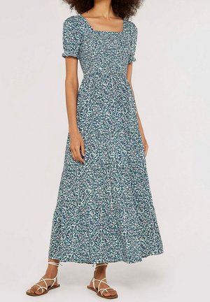 Femme portant une longue robe fleurie verte et blanche avec des manches courtes bouffantes et un décolleté carré, associée à des sandales plates blanches à lanières.