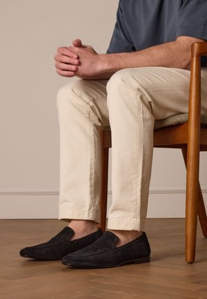 Black suede slip-on shoes with a pointed toe and elastic side accents, worn with light-colored, tailored chinos, seated on a wooden chair.