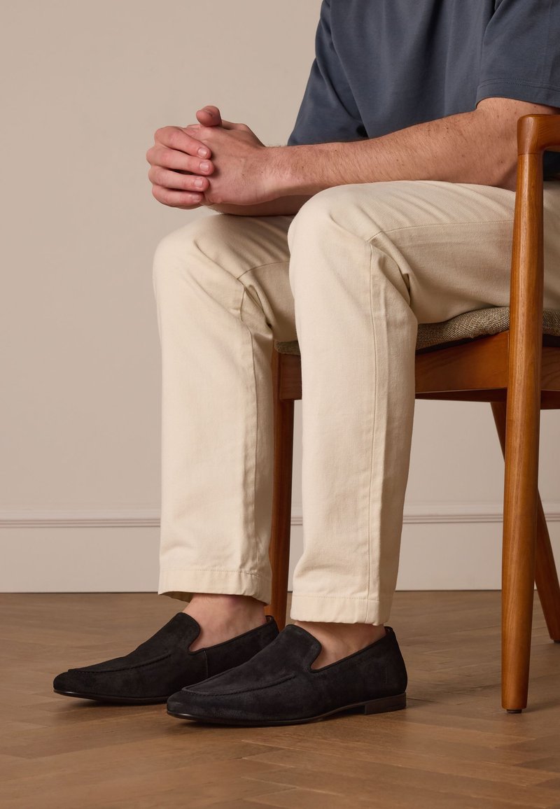 Zwarte suède instapchoenen met een spitse neus en elastische zijkanten, gedragen bij lichtgekleurde, op maat gemaakte chinos, zittend op een houten stoel.