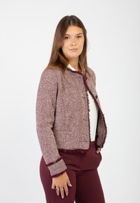 Blazer bordeaux in tessuto texturizzato con motivo a spina di pesce, bordi sfilacciati e chiusura a bottoni. Abbinato a un top bianco e pantaloni bordeaux coordinati.