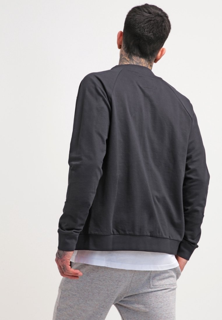 Homme au cou et aux mains tatoués, portant une chemise noire à manches longues par-dessus un t-shirt blanc et un pantalon gris clair, vu de dos sur un fond uni.