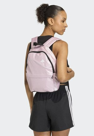 Frau, die einen hellrosa Rucksack mit schwarzen Reißverschlüssen, ein schwarzes ärmelloses Oberteil und schwarze Shorts mit weißen Streifen trägt, seitlich steht.