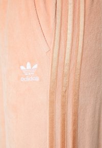 Narůžovělé sametové tepláky s elegantním designem. Obsahují tři béžové pruhy, boční kapsy a vyšitý bílý logo Adidas.