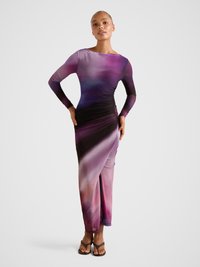 Moteris pozuoja dėvėdama ilgą, aptemptą suknelę su violetiniu ir rožiniu gradientu, ilgomis rankovėmis ir šoninėmis raukėmis, avinti rudus atvirus sandalų batus.