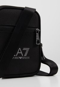 Borsa a tracolla nera in tessuto, con una texture liscia, caratterizzata da una cerniera frontale, logo argentato "A7" e brand "Emporio Armani", e una tracolla regolabile.