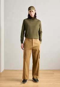 Man som bär en olivgrön polotröja, bruna cargobyxor, mörka sneakers och en olivgrön keps som står på ett trädkigt golv mot en enfärgad ljus vägg.