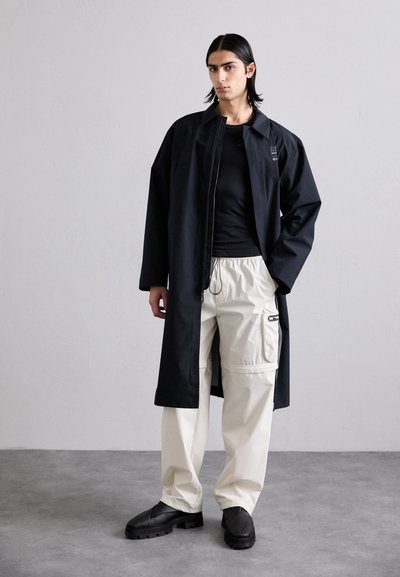 Casaco longo preto com um corte relaxado e zíper, combinado com calças cargo em creme com bolsos laterais. Calçado: sapatos pretos chunky com sulcos.