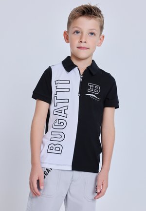 Ragazzo giovane in piedi, indossa una polo Bugatti bianca e nera e pantaloncini grigio chiaro con uno sfondo neutro.