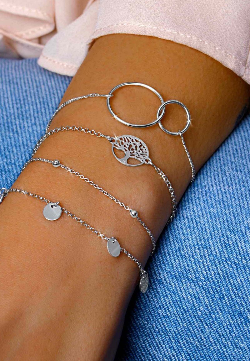 Conjunto de pulseras de plata que presenta anillos entrelazados, un diseño de árbol de la vida y charms redondos, todo sobre una muñeca bronceada y sobre tela de mezclilla.