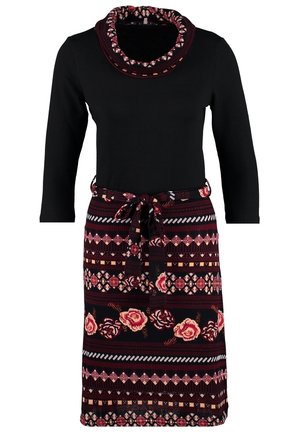 Kvinde iført sort top med 3/4-ærmer, mønstret blomsteret nederdel med bælte, sorte strømpebukser, beige ankelstøvler, holder en brun hat i venstre hånd.