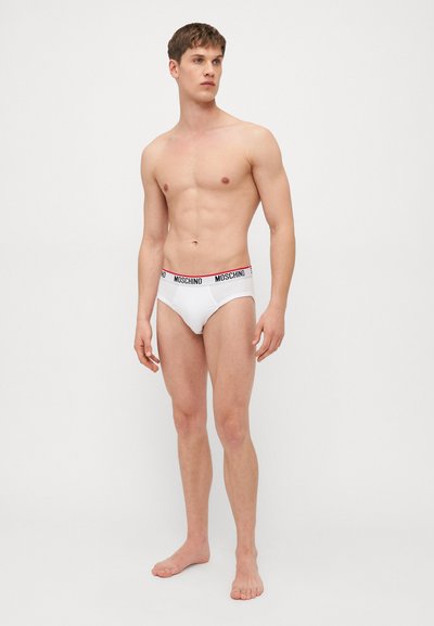 White men's briefs with a black and red Moschino waistband, featuring a smooth texture and a classic brief shape, displayed on a model.