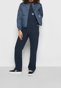 Granatowa, pikowana bomberka na granatowych, paskowanych ogrodniczkach z guzikami. Połączona z czarnymi trampkami na białym tle.