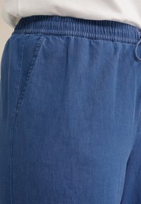 Mėlynos džinsinės kelnės jogger stiliaus su elastinga juosta, šoninėmis kišenėmis ir minkšta tekstūra. Audinys atrodo lengvas ir kasdienis.