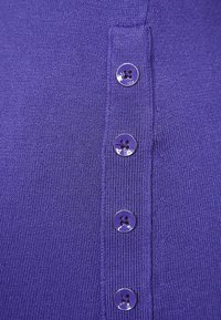 Fialový pletený oděv s pěti knoflíkovou légou, která obsahuje kulaté fialové knoflíky s výřezy ve tvaru květin. Hladká textura a přiléhavý design.