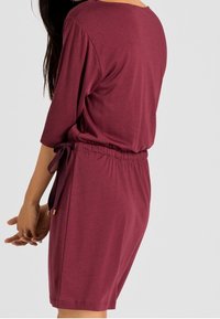 Burgundy dress with 3/4 sleeves, a relaxed fit, an elastic waistband, and a adjustable tie detail at the waist. Soft fabric texture.