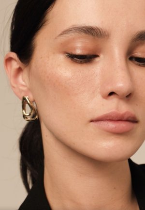 Mujer de cabello oscuro que lleva un moderno pendiente de aro dorado, mirando hacia abajo, con piel tersa y maquillaje neutro.